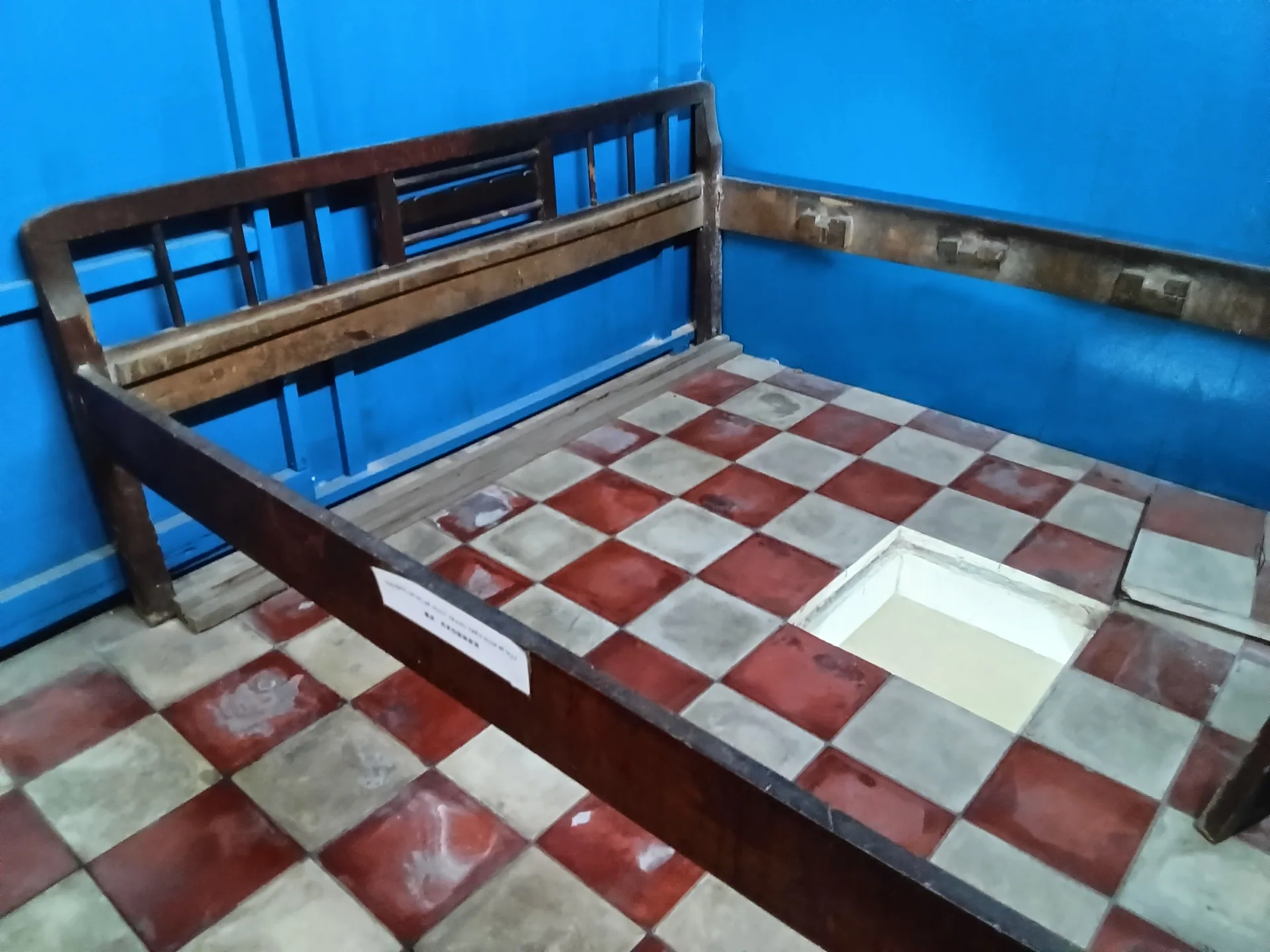 Khung giường và nắp hầm bí mật mở trên nền gạch trong căn phòng màu xanh.