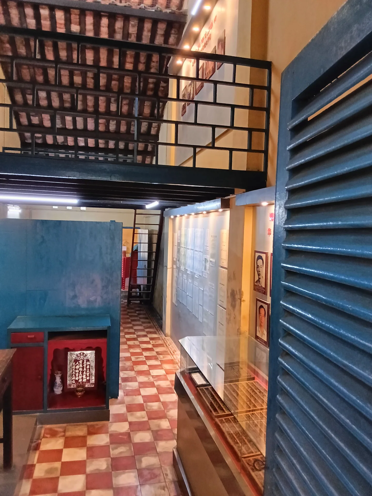 Góc nhìn trong nhà di tích với cửa xanh, nền gạch caro và phần mái, gác gỗ phía trên.